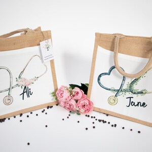 K&ouml;nnte beinhalten: Zwei personalisierte Jute-Tragetaschen mit wei&szlig;en Stoffeins&auml;tzen. Die Taschen zeigen ein Stethoskop mit einem Herzen und einem Blumendesign. Eine Tasche sagt "Ali" und die andere sagt "Jane".