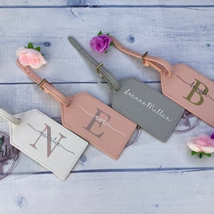 Pu&ograve; includere: Quattro etichette per bagagli personalizzate con cinturini in pelle. Le etichette sono di diversi colori: bianco, rosa, grigio e rosa chiaro. Ogni etichetta ha un'iniziale dorata e un nome o un'iniziale scritto a mano. Le etichette sono su uno sfondo di legno bianco.