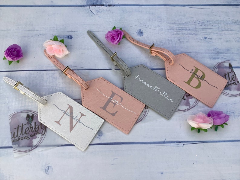 Pu&ograve; includere: Quattro etichette per bagagli personalizzate con cinturini in pelle. Le etichette sono di diversi colori: bianco, rosa, grigio e rosa chiaro. Ogni etichetta ha un'iniziale dorata e un nome o un'iniziale scritto a mano. Le etichette sono su uno sfondo di legno bianco.