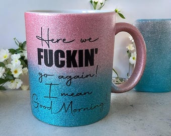 Può includere: Tazza in ceramica glitterata, sfumata dal rosa al turchese, con la scritta "Here we FUCKIN' go again! I mean Good Morning!" in nero. La tazza ha un grande manico e un interno bianco. Un'altra tazza turchese è sullo sfondo.