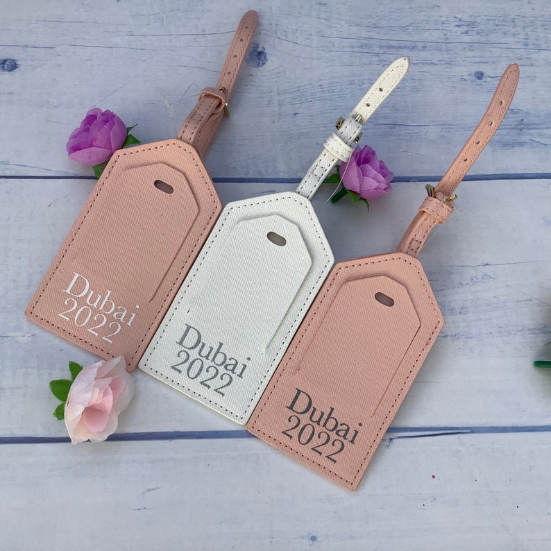 Puede incluir: Tres etiquetas de equipaje de material similar al cuero en color rosa y blanco. Cada etiqueta tiene una correa blanca y dice "Dubai 2022" en letras blancas.