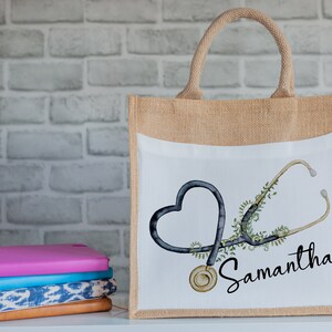 K&ouml;nnte beinhalten: Eine braune Jute-Tragetasche mit einem wei&szlig;en Canvas-Einsatz, der ein schwarzes und gr&uuml;nes Stethoskop in Herzform und den Namen Samantha zeigt.