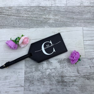 Pu&ograve; includere: Etichetta per bagagli in pelle nera con una lettera C bianca e la parola "Caitlin" scritta in corsivo. L'etichetta &egrave; su uno sfondo in legno chiaro con fiori rosa e viola.