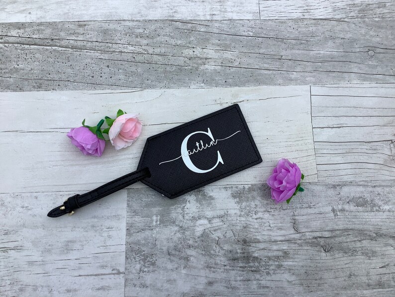 Pu&ograve; includere: Etichetta per bagagli in pelle nera con una lettera C bianca e la parola "Caitlin" scritta in corsivo. L'etichetta &egrave; su uno sfondo in legno chiaro con fiori rosa e viola.