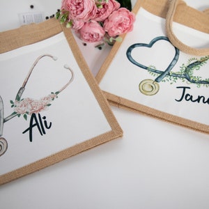 K&ouml;nnte beinhalten: Zwei personalisierte Jute-Tragetaschen mit einem Stethoskop und einem Blumendesign. Die Tasche links tr&auml;gt den Namen "Ali" und die Tasche rechts den Namen "Jane".