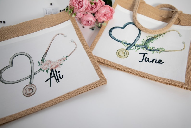 K&ouml;nnte beinhalten: Zwei personalisierte Jute-Tragetaschen mit einem Stethoskop und einem Blumendesign. Die Tasche links tr&auml;gt den Namen "Ali" und die Tasche rechts den Namen "Jane".