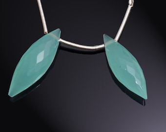 May include: Two faceted, teardrop-shaped, pale teal gemstones strung on a silver chain. The gemstones have a pointed tip and a series of angled facets that catch the light. The background is a dark, reflective surface.