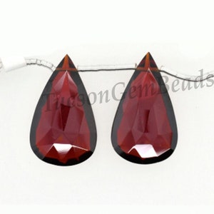 May include: Two pear-shaped red gemstone beads with faceted surfaces. The beads are strung on a silver wire.
