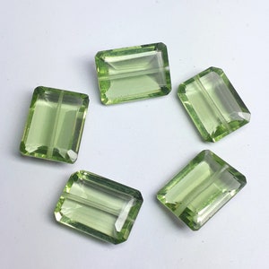 Puede incluir: Cinco gemas rectangulares facetadas de color verde claro dispuestas sobre una superficie blanca. Cada gema tiene forma rectangular con bordes cortados, creando un efecto brillante. Las gemas son probablemente para la fabricación de joyas.