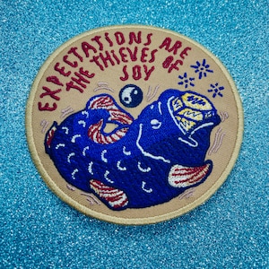 Könnte beinhalten: Ein beigefarbener gestickter Aufnäher mit einem blauen Fisch mit roten und weißen Streifen. Der Fisch verschlingt ein Yin-Yang-Symbol. Der Text auf dem Aufnäher lautet "Expectations are the thieves of joy".