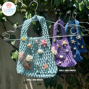 May include: Three handmade macrame shell bags in shades of blue and purple, adorned with seashells and starfish. The bags are hanging on metal hangers. The text "SHELL-BAG-GREEN" and "SHELL-BAG-PURPLE" are visible. The bags are a beach decor item.