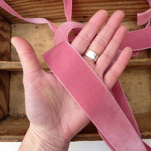 Puede incluir: Una mano sosteniendo una cinta de terciopelo rosa polvoriento. La cinta tiene aproximadamente 2,5 cm de ancho y una textura suave y aterciopelada.