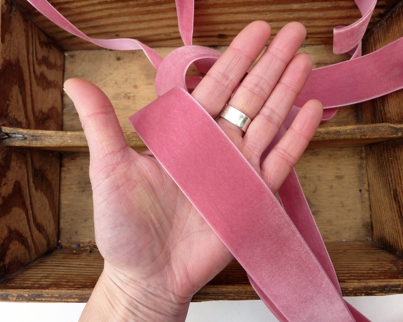 Puede incluir: Una mano sosteniendo una cinta de terciopelo rosa polvoriento. La cinta tiene aproximadamente 2,5 cm de ancho y una textura suave y aterciopelada.