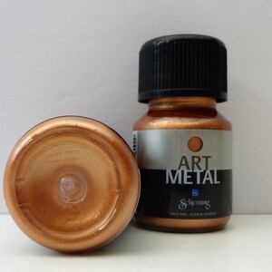 K&ouml;nnte beinhalten: Zwei Flaschen kupferfarbener Metallic-Farbe. Die Flasche rechts hat einen schwarzen Deckel und ein Etikett mit der Aufschrift "Art Metal" und darunter dem Markennamen "Schjerning". Die Flasche links steht auf dem Kopf und zeigt die kupferfarbene Farbe.