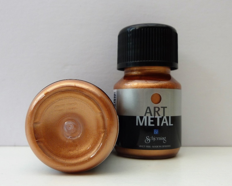 K&ouml;nnte beinhalten: Zwei Flaschen kupferfarbener Metallic-Farbe. Die Flasche rechts hat einen schwarzen Deckel und ein Etikett mit der Aufschrift "Art Metal" und darunter dem Markennamen "Schjerning". Die Flasche links steht auf dem Kopf und zeigt die kupferfarbene Farbe.