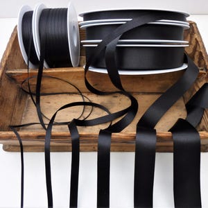 May include: Black satin ribbon spools displayed in a rustic wooden tray. The ribbon is unwound in several lengths, showcasing its smooth texture and sheen. The spools are white with black ribbon.