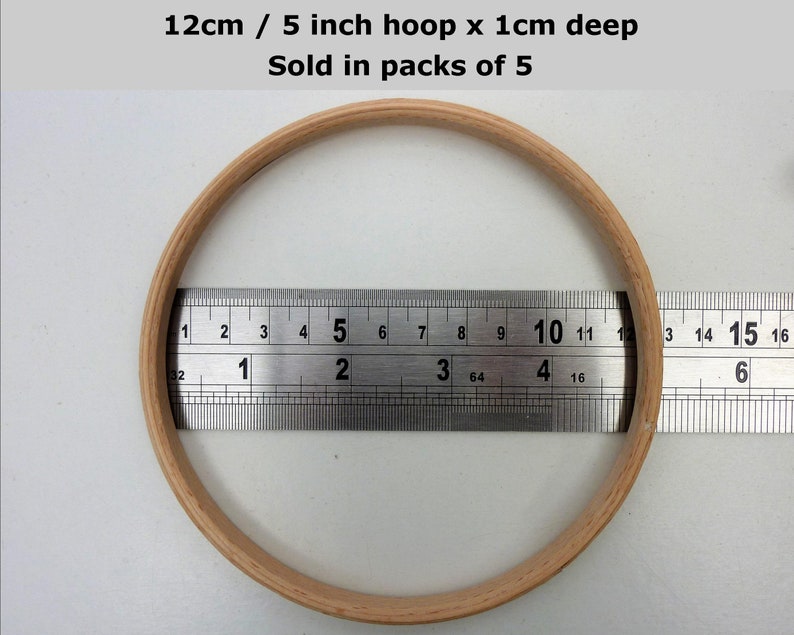 May include: A wooden embroidery hoop with a diameter of 5 inches and a depth of 1 cm. The hoop is being measured with a ruler. The text "12cm / 5 inch hoop x 1cm deep Sold in packs of 5" is visible in the image.