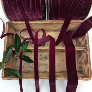 May include: Two spools of deep burgundy velvet ribbon are positioned above a rectangular wooden crate. Several lengths of the dark red ribbon drape down from the spools into the crate, accompanied by a small green leafy branch on the left side.