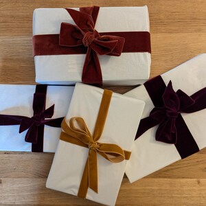 Puede incluir: Cuatro cajas de regalo blancas envueltas con cintas de terciopelo de diferentes colores. Las cajas est&aacute;n sobre una superficie de madera. Las cintas son burdeos, doradas y moradas.