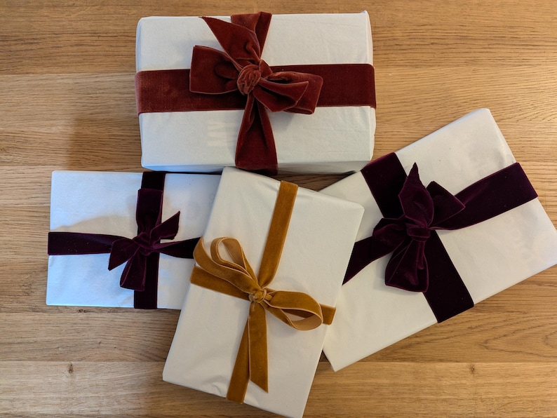 Puede incluir: Cuatro cajas de regalo blancas envueltas con cintas de terciopelo de diferentes colores. Las cajas est&aacute;n sobre una superficie de madera. Las cintas son burdeos, doradas y moradas.