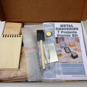 K&ouml;nnte beinhalten: Ein Metallpr&auml;ge-Starter-Set mit 7 Projekten. Das Set enth&auml;lt einen Holzrahmen, ein Notizbuch, einen Pinsel, einen Stift, eine Metallplatte und eine Brosch&uuml;re mit Anweisungen. Das Set ist in einer wei&szlig;en Box mit einem braunen Pappdeckel verpackt.