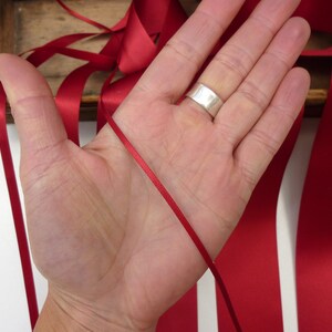 Puede incluir: Una mano sosteniendo una fina cinta de raso roja, con cintas rojas m&aacute;s anchas en el fondo. La cinta es de un color rojo intenso y rico. Un anillo plateado est&aacute; en la mano. Se ve una caja de madera.