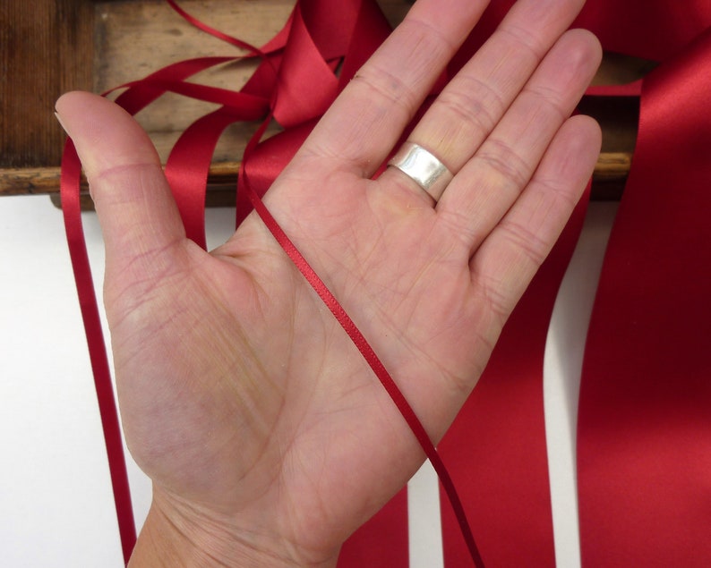 Puede incluir: Una mano sosteniendo una fina cinta de raso roja, con cintas rojas m&aacute;s anchas en el fondo. La cinta es de un color rojo intenso y rico. Un anillo plateado est&aacute; en la mano. Se ve una caja de madera.