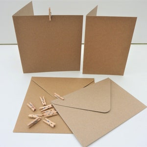 May include: Kraft paper greeting cards and envelopes with small wooden pegs. The cards are folded and propped up, while the envelopes and pegs are arranged on a white surface. The paper has a natural, earthy tone.