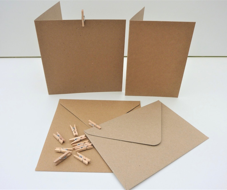 May include: Kraft paper greeting cards and envelopes with small wooden pegs. The cards are folded and propped up, while the envelopes and pegs are arranged on a white surface. The paper has a natural, earthy tone.