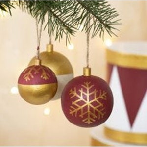 May include: Three Christmas ornaments with a gold and burgundy color scheme. The ornaments are decorated with a snowflake design in gold paint.
