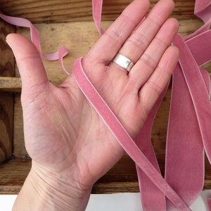 Puede incluir: Primer plano de una mano sosteniendo varios hilos de cinta de terciopelo rosa. La cinta es suave y lujosa, y ser&iacute;a perfecta para agregar un toque de elegancia a cualquier proyecto.