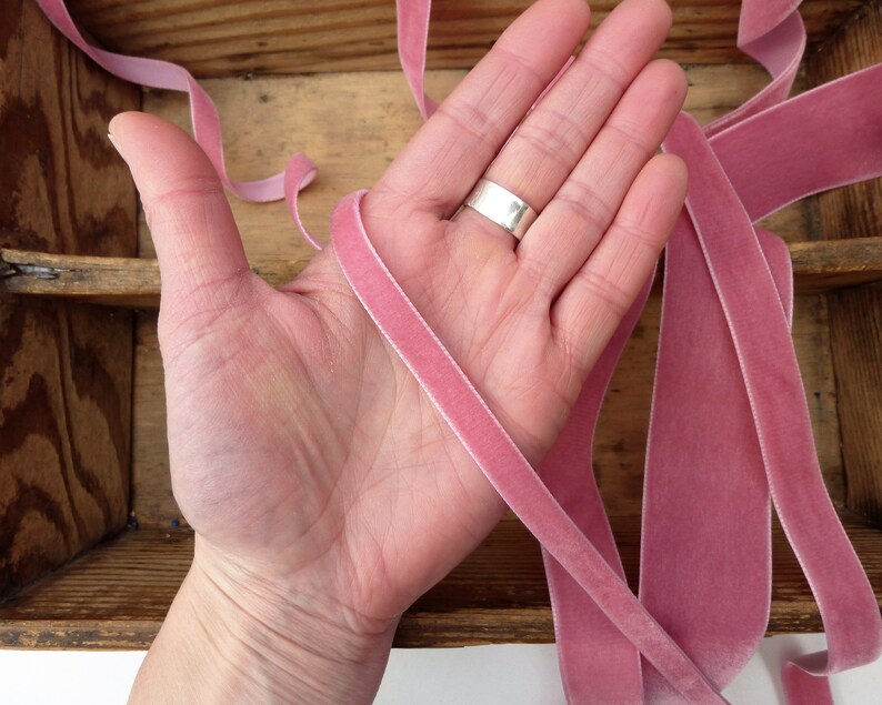 Puede incluir: Primer plano de una mano sosteniendo varios hilos de cinta de terciopelo rosa. La cinta es suave y lujosa, y ser&iacute;a perfecta para agregar un toque de elegancia a cualquier proyecto.