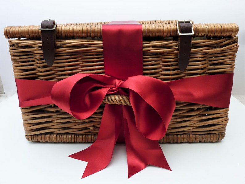Puede incluir: Cesta de picnic de mimbre con tapa con bisagras, asegurada con correas y hebillas de cuero marr&oacute;n. Una gran cinta de raso rojo vibrante est&aacute; atada alrededor de la cesta, formando un lazo decorativo. La cesta est&aacute; vac&iacute;a.