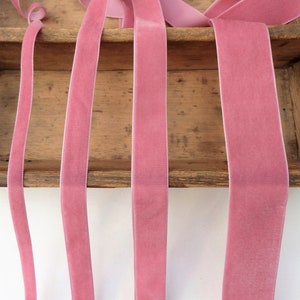 Puede incluir: Cinco carretes de cinta de terciopelo rosa de diferentes anchos. La cinta se muestra en un estante de madera.