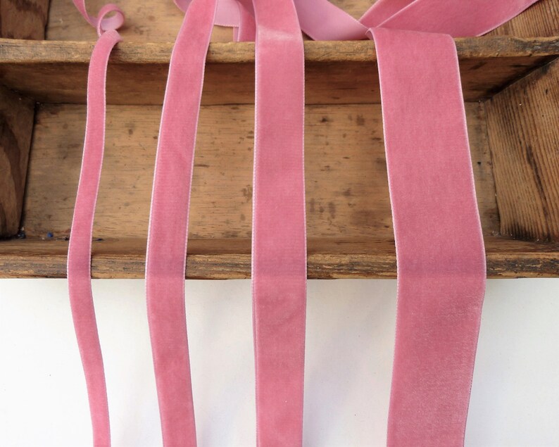 Puede incluir: Cinco carretes de cinta de terciopelo rosa de diferentes anchos. La cinta se muestra en un estante de madera.