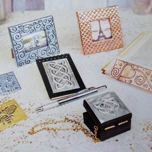 K&ouml;nnte beinhalten: Eine Sammlung von sieben Metallbilderrahmen, Boxen und einem Notizbuch. Die Rahmen und Boxen sind mit gepr&auml;gten Designs verziert, darunter Spiralen, Herzen und keltische Knoten. Das Notizbuch hat einen schwarzen Lederumschlag mit einem keltischen Knotendesign. Die Artikel sind auf einer wei&szlig;en Spitzentischdecke angeordnet.