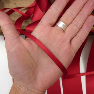 Puede incluir: Cinta de raso rojo carmes&iacute;, un material de manualidades vers&aacute;til, se muestra. La cinta, de aproximadamente 1,3 cm de ancho, se sostiene en una mano, mostrando su textura suave y color vibrante. Ideal para proyectos creativos.