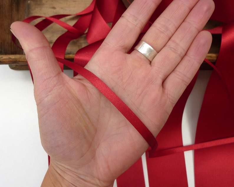 Puede incluir: Cinta de raso rojo carmes&iacute;, un material de manualidades vers&aacute;til, se muestra. La cinta, de aproximadamente 1,3 cm de ancho, se sostiene en una mano, mostrando su textura suave y color vibrante. Ideal para proyectos creativos.