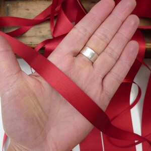 Puede incluir: Una mano sostiene un trozo de cinta de raso roja. La cinta es lisa y tiene un ligero brillo. Otras longitudes de la misma cinta son visibles en el fondo, junto con una caja de madera.
