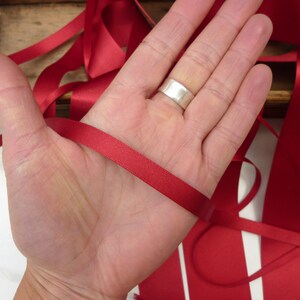 Puede incluir: Una mano sosteniendo un trozo de cinta de raso rojo brillante. La cinta es lisa y tiene un ligero brillo. Otras longitudes de cinta son visibles en el fondo, junto con una caja de madera.