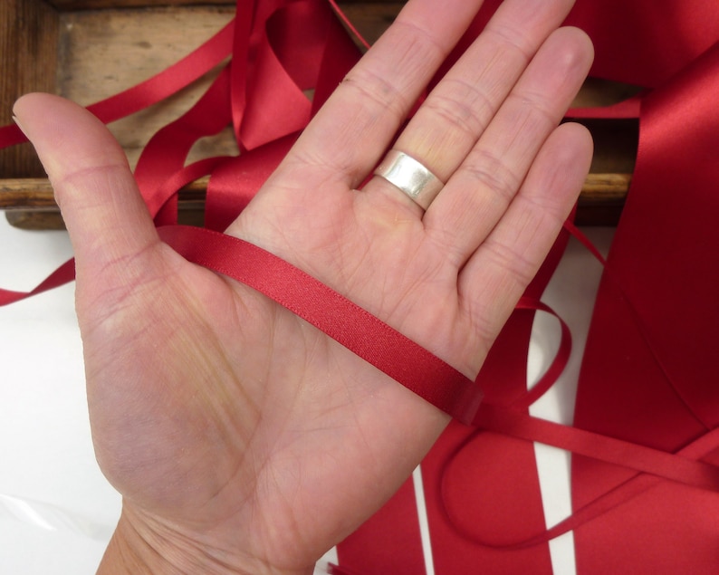Puede incluir: Una mano sosteniendo un trozo de cinta de raso rojo brillante. La cinta es lisa y tiene un ligero brillo. Otras longitudes de cinta son visibles en el fondo, junto con una caja de madera.