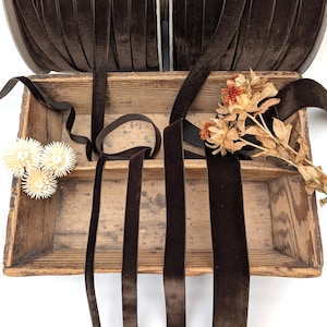 Puede incluir: Un carrete de cinta de terciopelo marrón oscuro con varias longitudes de cinta dispuestas en una caja de madera rectangular. La caja contiene tres pequeñas flores secas blancas a la izquierda y un ramo de flores secas naranjas y beige a la derecha.
