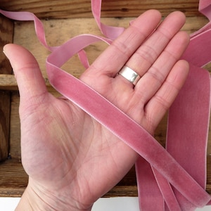 Puede incluir: Primer plano de una mano sosteniendo dos largos de cinta de terciopelo rosa polvoriento. La cinta es suave y lujosa, perfecta para agregar un toque de elegancia a cualquier proyecto.