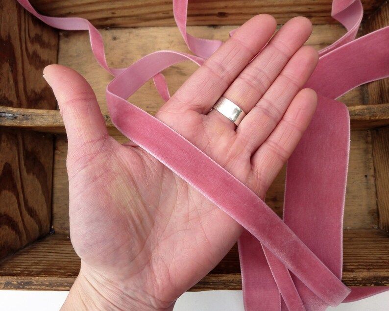 Puede incluir: Primer plano de una mano sosteniendo dos largos de cinta de terciopelo rosa polvoriento. La cinta es suave y lujosa, perfecta para agregar un toque de elegancia a cualquier proyecto.