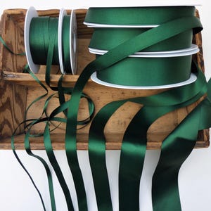 May include: An assortment of dark green satin ribbon spools and loose ribbon strands. The spools are displayed in a rustic wooden tray. The ribbon has a glossy finish and is ideal for crafts, gift wrapping, or decorative purposes.