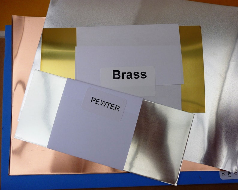 K&ouml;nnte beinhalten: Nahaufnahme von vier Metallblechen mit den Beschriftungen "Brass", "Pewter" und "Copper". Die Bleche sind &uuml;bereinander gestapelt, wobei das Messingblech oben liegt, gefolgt vom Zinnblech, dem Kupferblech und einem blauen Blech unten.
