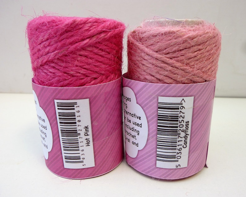 May include: Two spools of pink jute twine, one a darker hot pink and the other a lighter candy floss pink. Both spools have barcodes and product information labels.