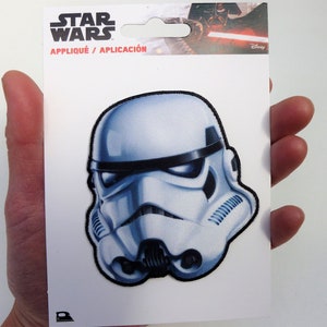 May include: A white and black Star Wars iron-on patch featuring a Stormtrooper helmet. The patch is on a white card with the text "Star Wars" and "Appliqué / Aplicación".