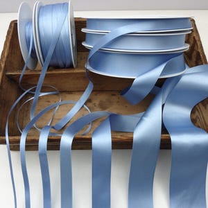 May include: Several spools of light blue satin ribbon are displayed in a rustic wooden tray. The ribbon is unwound from the spools, with lengths of the ribbon cascading down. The ribbon has a smooth, shiny texture, perfect for crafts and gift wrapping.