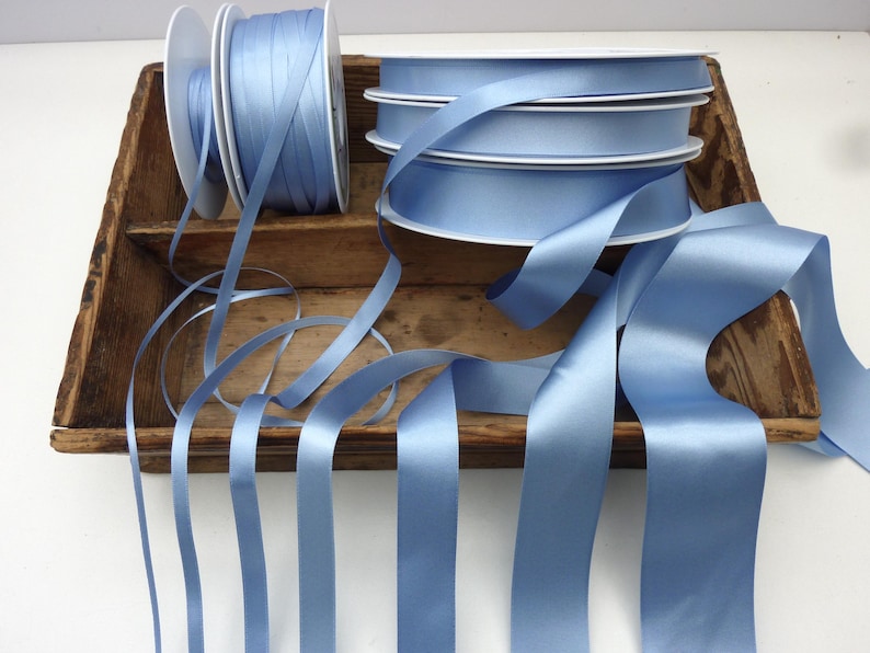 May include: Several spools of light blue satin ribbon are displayed in a rustic wooden tray. The ribbon is unwound from the spools, with lengths of the ribbon cascading down. The ribbon has a smooth, shiny texture, perfect for crafts and gift wrapping.